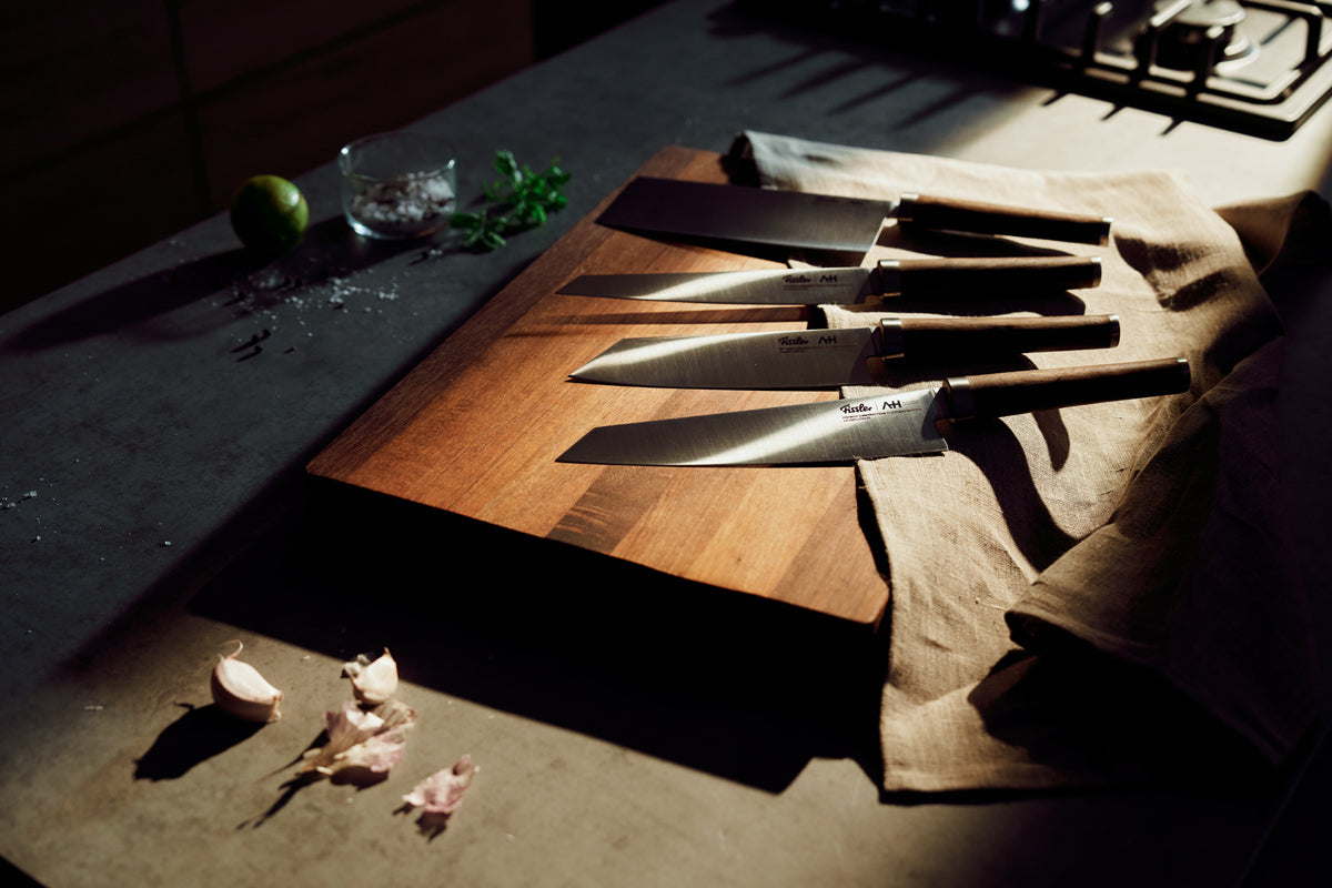Fissler PrimeCut Santokumesser 17cm auf Holzbrett mit Knoblauch, Kalk und Sonnenlicht - moderne deutsche Qualität trifft auf zeitloses Design.