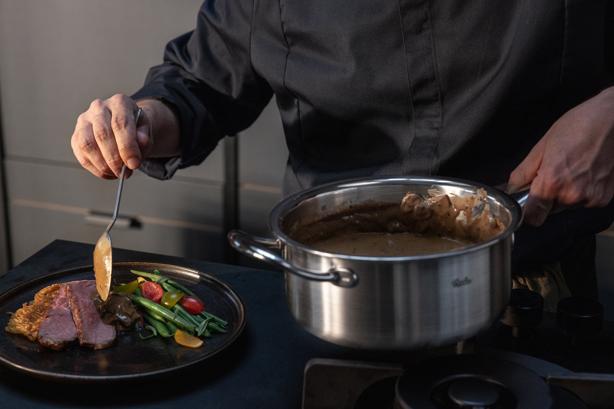 Ein Koch in dunkler Kleidung gießt Soße aus einem Fissler-Edelstahltopf über in Scheiben geschnittenes Fleisch, das neben farbenfrohem Gemüse auf einem schwarzen Teller liegt, und demonstriert so die Präzision und Qualität des 3-teiligen Fissler M5 Pro-Ply®-Sets mit Metalldeckel in einer modernen Küche. Die Szene unterstreicht Fisslers Tradition deutscher Ingenieurskunst, langlebiger Materialien und Innovation für zeitlose Kochleistung.