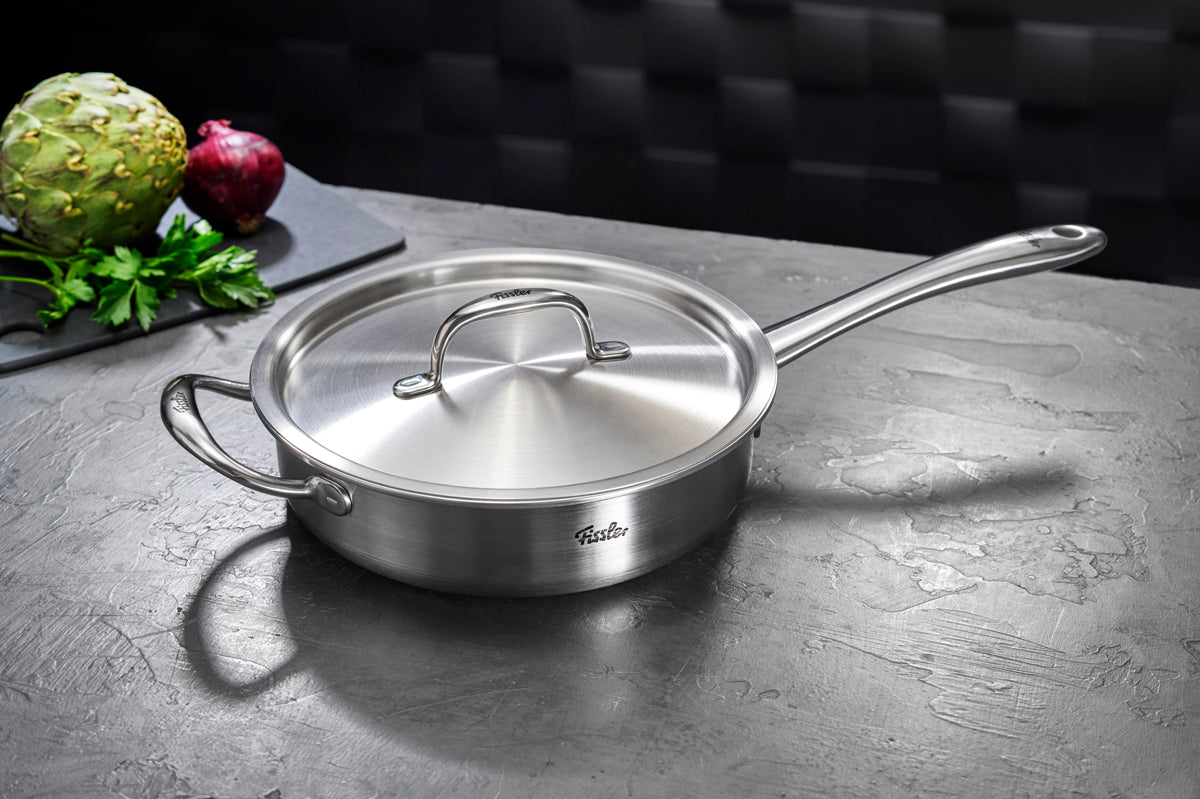 Fissler M5 Pro Ply® 4-teilige Sautierpfanne mit Deckel auf einer grauen Arbeitsplatte, im Hintergrund frisches Gemüse auf einem schwarzen Brett.