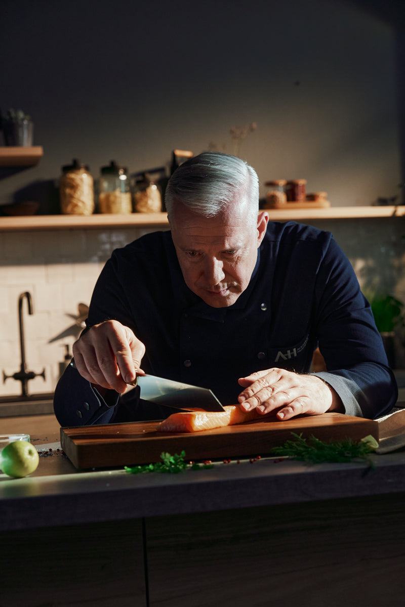 Ein Koch verwendet den Fissler PrimeCut Santokumesser 17cm zum Schneiden von Lachs, mit eleganten Gläsern und Utensilien auf modernen Küchenregalen.