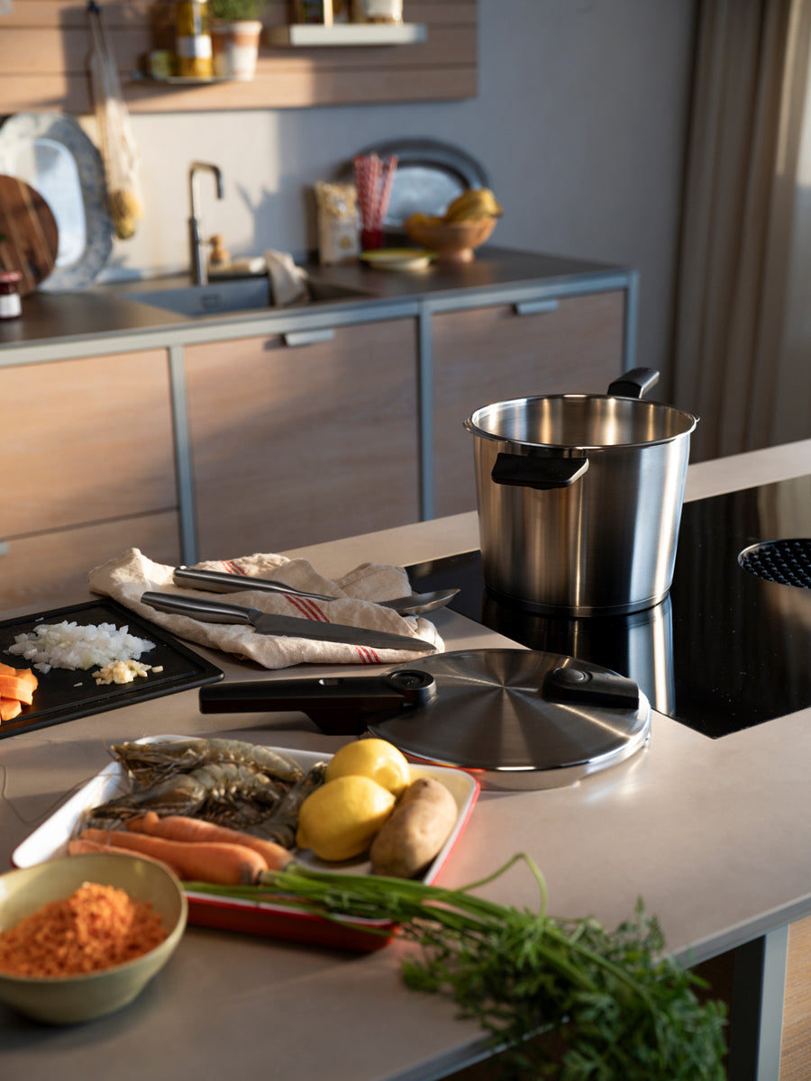 In einer modernen Küche steht das 2-teilige Fissler Vitaquick® Premium-Schnellkochtopfset mit SKT Buch im Mittelpunkt – fachmännisch in Deutschland gefertigt und bekannt für seine Präzisionstechnik, Langlebigkeit und sein zeitloses Design. Edelstahltöpfe und ein passender Topfdeckel sind auf der Arbeitsplatte neben einem Induktionsherd von frisch geschnittenem Gemüse, Zitronen und rohen Garnelen umgeben. Das ikonische Kochgeschirr von Fissler fügt sich nahtlos in den modernen Raum ein, während im