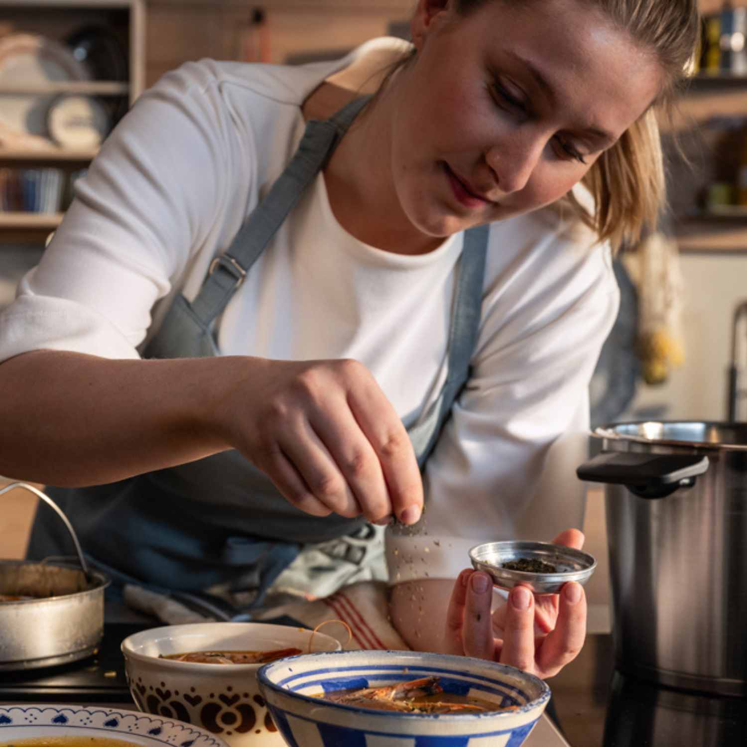 Eine Person in einer Schürze streut in einer Küche Gewürze über eine Schüssel mit Essen, umgeben von hochwertigen Fissler-Edelstahltöpfen und -Kochgeschirr. Die Szene spiegelt Fisslers Tradition von Präzisionstechnik, zeitlosem Design und Innovation „Made in Germany“ wider.