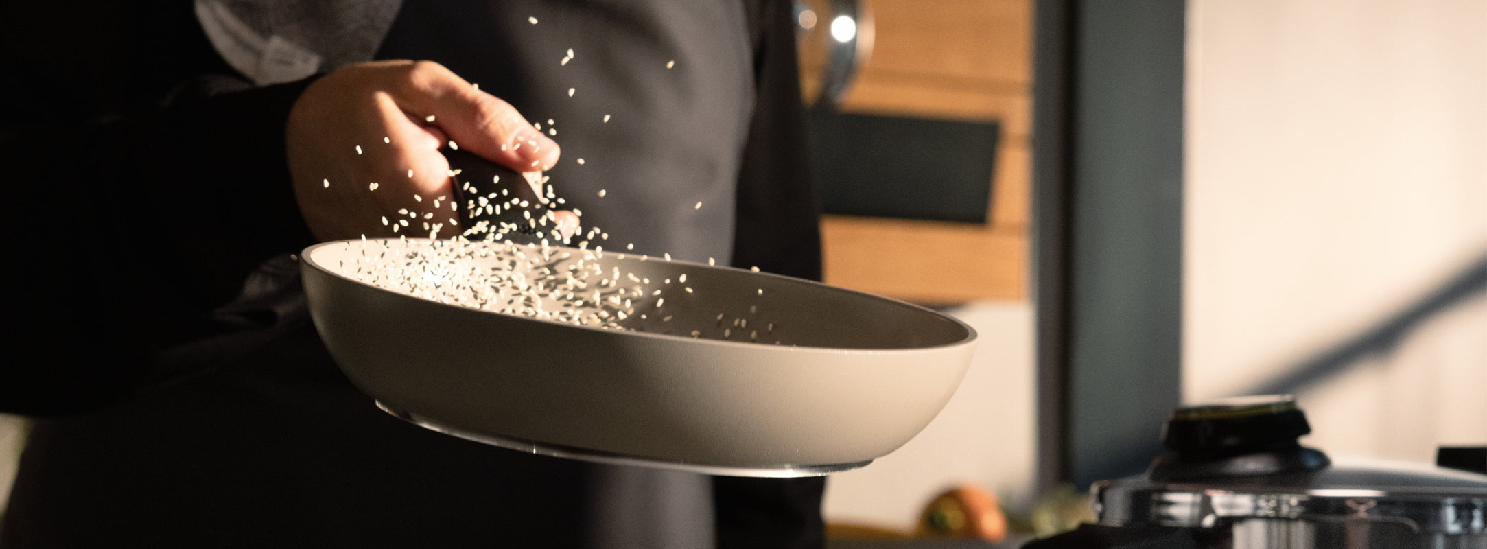 Eine Person schwenkt gekonnt Reis in einer beigen Fissler-Pfanne, Körner in der Luft, aufgenommen in einer hellen Küche mit weichem, unscharfem Hintergrund.