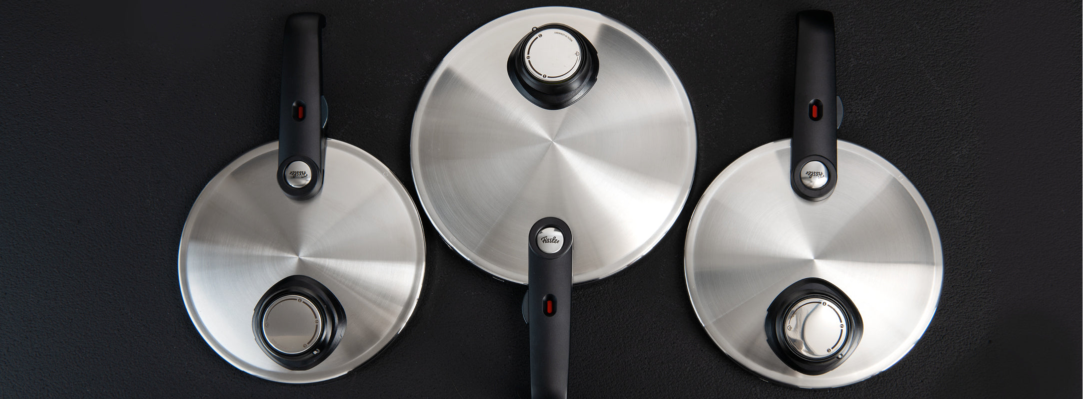 Three Fissler stainless steel pot lids with characteristic black knobs, arranged on a dark surface - precision meets timeless design.