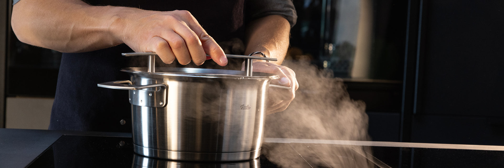 Eine Person hebt den Deckel von einem Fissler-Edelstahltopf, der in Deutschland hergestellt wird und aus dessen Präzisionsdesign elegant Dampf aufsteigt.