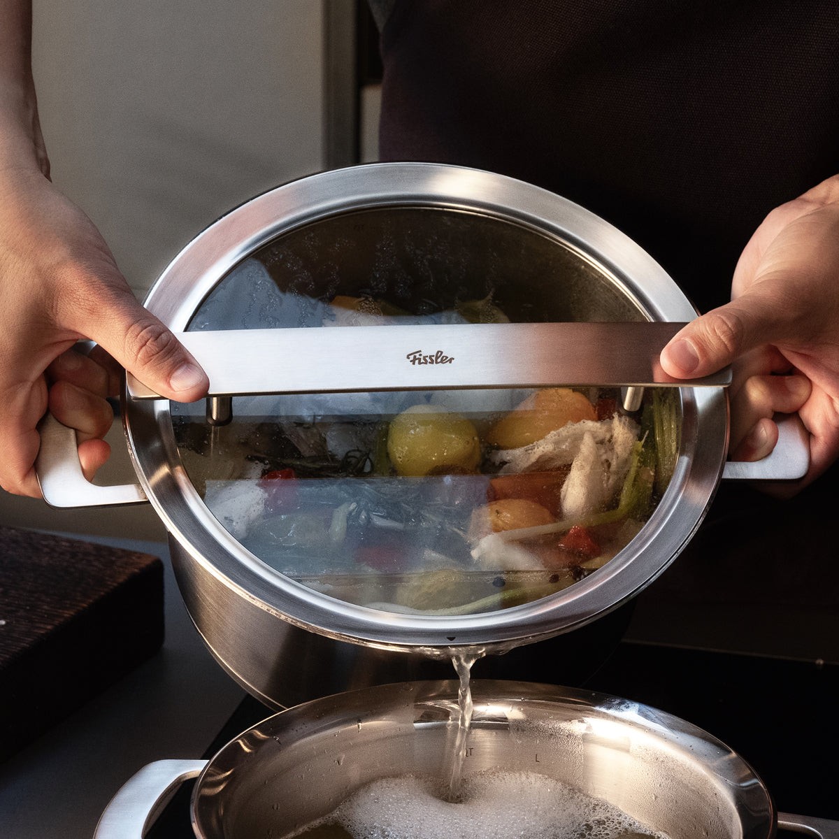 Ein Fissler-Edelstahltopf mit Glasdeckel und eingebautem Sieb lässt Gemüse und Fleisch in einen weiteren hochwertigen Fissler-Topf abtropfen.