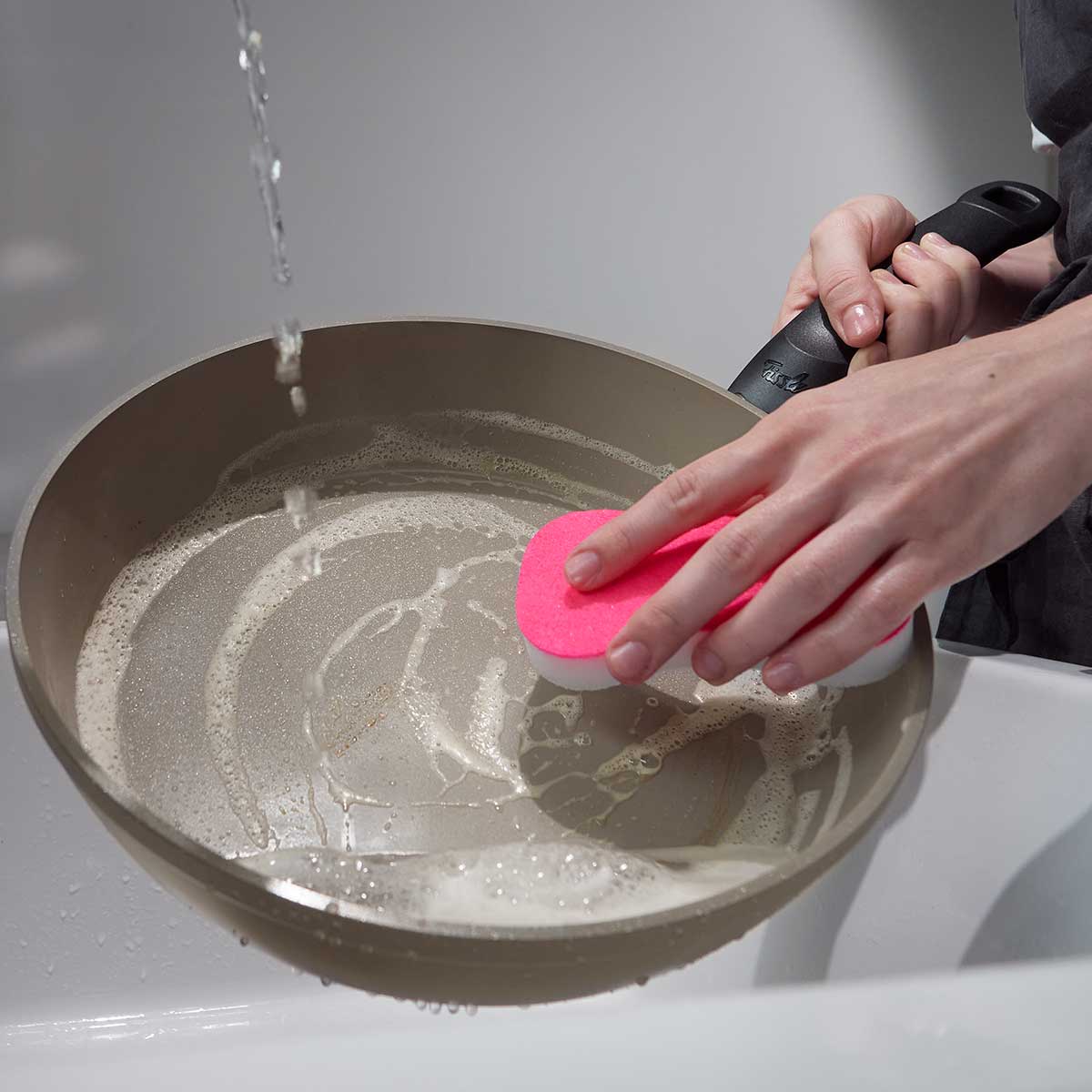 A person cleans a Fissler Ceratal® Comfort pan set (24 cm & 28 cm) with a pink sponge under running water, demonstrating German quality.
