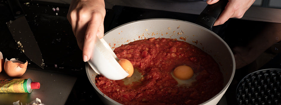 A chef cracks eggs into tomato sauce in a Fissler stainless steel pan; next to it are bowls, tomato paste, and chopped onions.