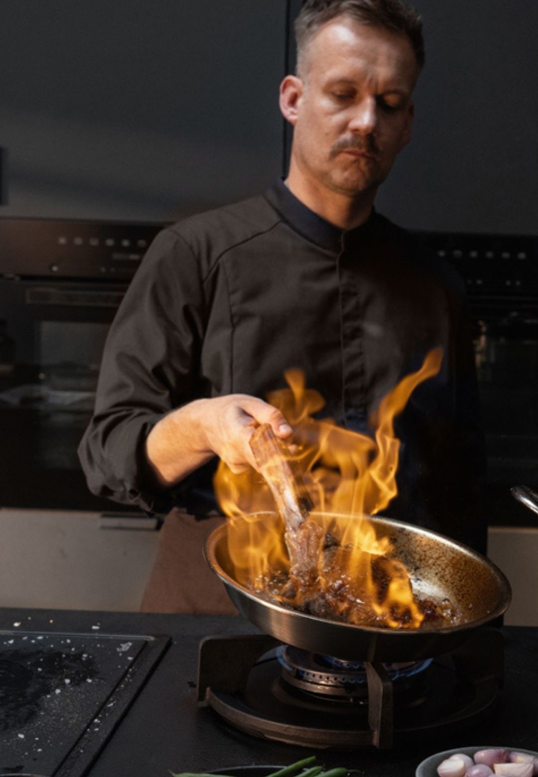 Ein Koch in dunkler Uniform brät Fleisch in einer hochwertigen Fissler-Pfanne über Gasflammen an; die Küchenkulisse ist sanft beleuchtet und raffiniert.