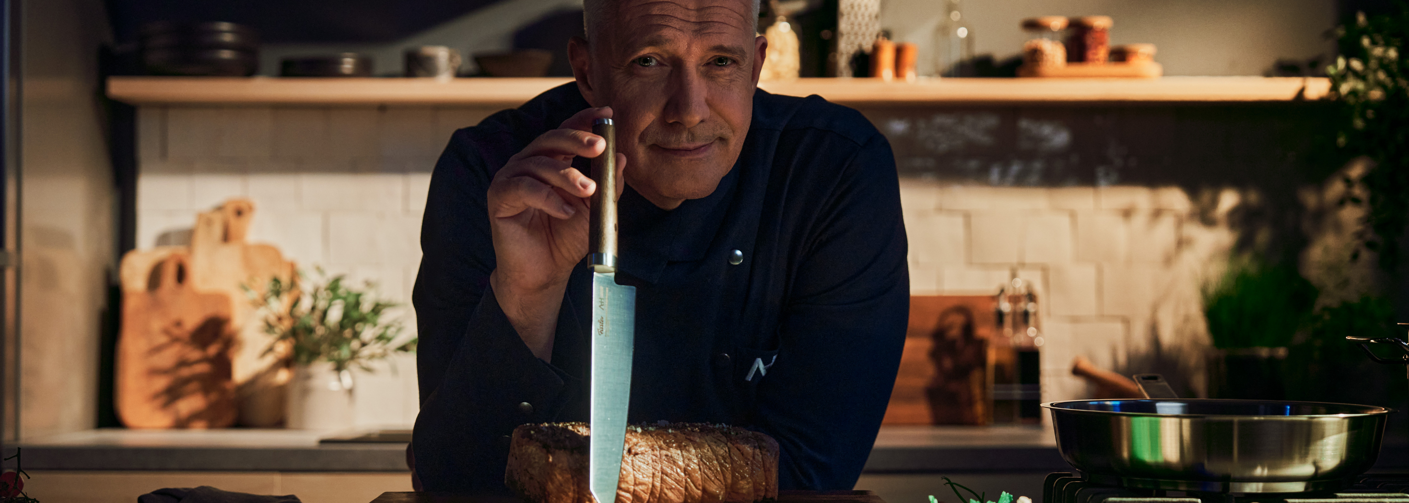 A chef in a dark kitchen holds a high-quality Fissler knife next to fresh bread on a wooden board, with Fissler cookware in the background.