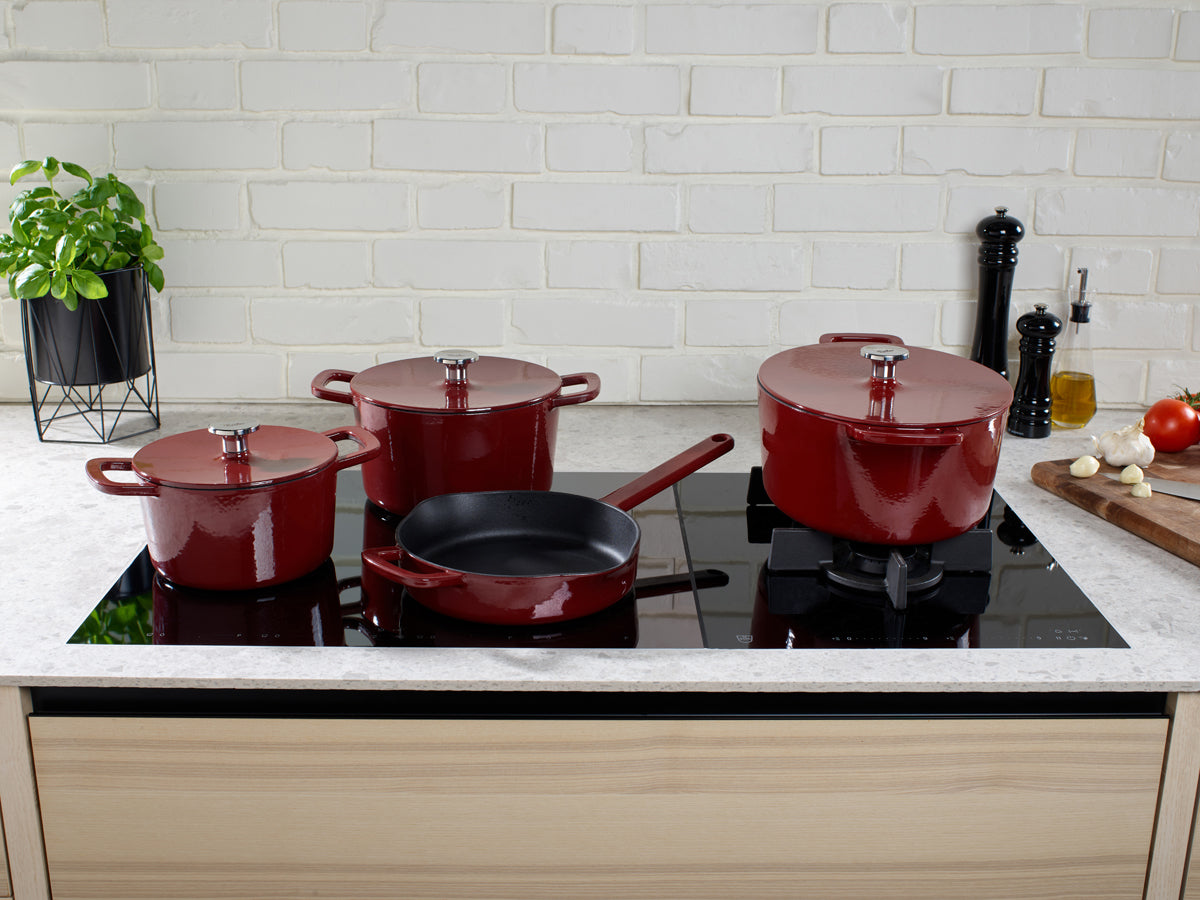 Vier rote Fissler Moments Gusseisen-Bräter mit Deckel auf Induktionsherd, umgeben von frischen Zutaten - Made in Germany.