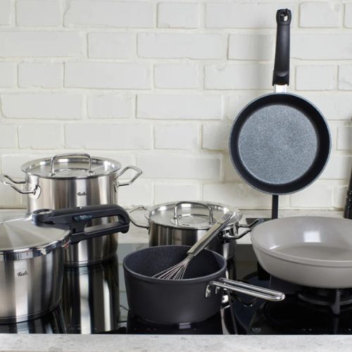Töpfe, Pfannen und ein Schneebesen aus Edelstahl von Fissler auf einer Herdplatte in einer modernen Küche mit weißem Backsteinsockel, die deutsche Qualität zeigt.