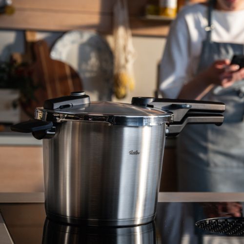 Ein Fissler Schnellkochtopf aus rostfreiem Stahl steht auf einer Herdplatte in einer eleganten Küche; ein verschwommener Koch in einer Schürze steht dahinter und hält ein Telefon.