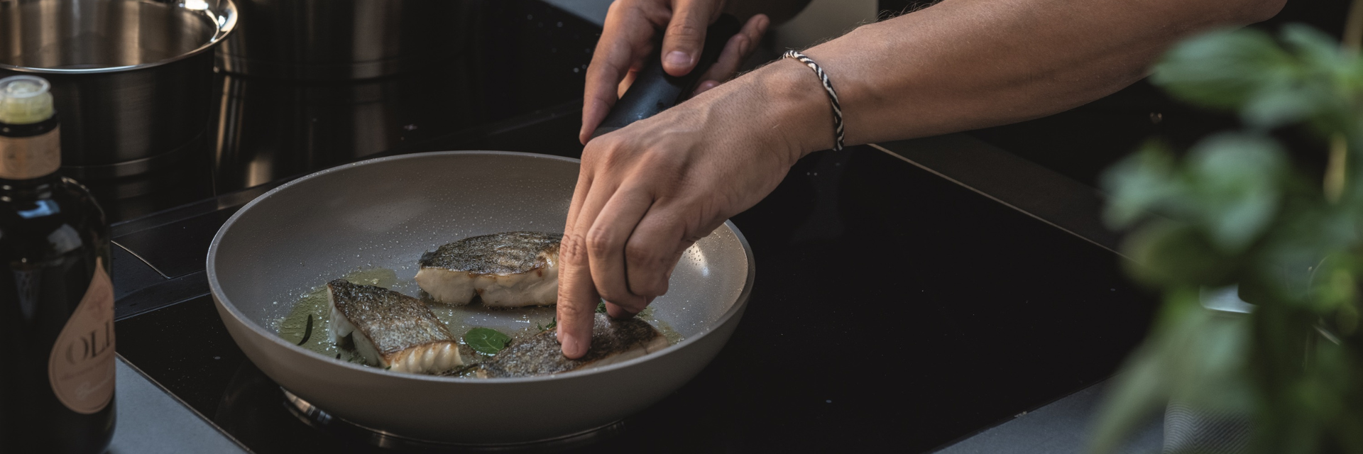 A person arranges fish fillets in a Fissler pan on the stove, with fresh herbs and a bottle next to the high-quality cookware.