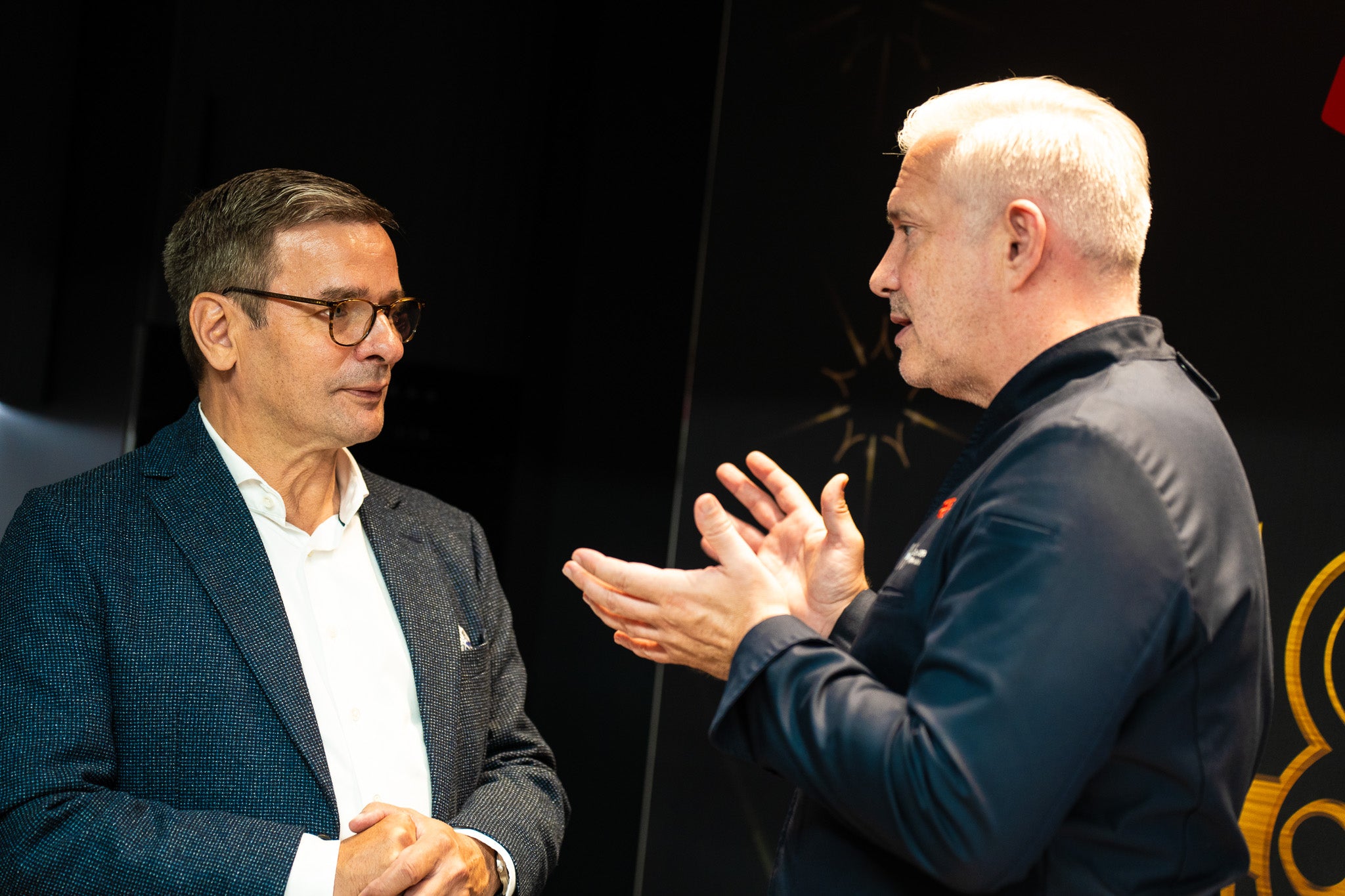 Two men discuss in a modern kitchen; high-quality Fissler stainless steel cookware sits on the counter next to them.
