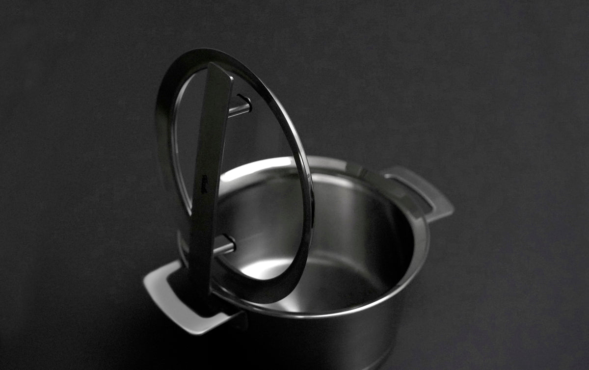 Fissler Phi Collection Edelstahltopf mit zwei Henkeln, teilweise abgedeckt durch einen 20 cm hohen Glasdeckel, steht vor einem dunklen Hintergrund.
