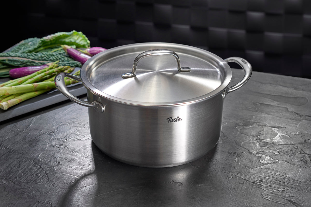 Das 4-teilige Fissler M5 Pro-Ply®-Topfset mit Metalldeckeln, gefertigt aus recyceltem Edelstahl „Made in Germany“, präsentiert sich mit robusten, ergonomischen Griffen auf einer dunklen Arbeitsplatte. Im Hintergrund sorgen frischer Spargel und Blattgemüse für Kochinspiration. Dieses hochwertige Set verkörpert Fisslers Tradition aus Präzision, Langlebigkeit und innovativem Design – optimal für Induktionsherde sowie vielseitiges Kochen.