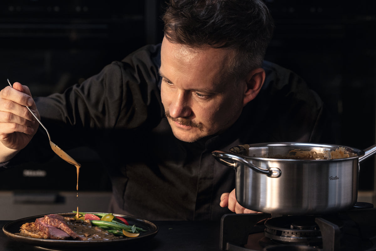 Ein Koch in schwarzer Jacke gießt mit einem Löffel aus recyceltem Edelstahl gekonnt Soße über ein auf einem Teller angerichtetes Gericht aus Gemüse und Fleisch. Neben dem Koch am Herd steht die Fissler M5 Pro-Ply® Stielkasserolle mit Metalldeckel – ein induktionsgeeigneter Topf, der für seine präzise Verarbeitung, außergewöhnliche Langlebigkeit und sein zeitloses deutsches Design bekannt ist.