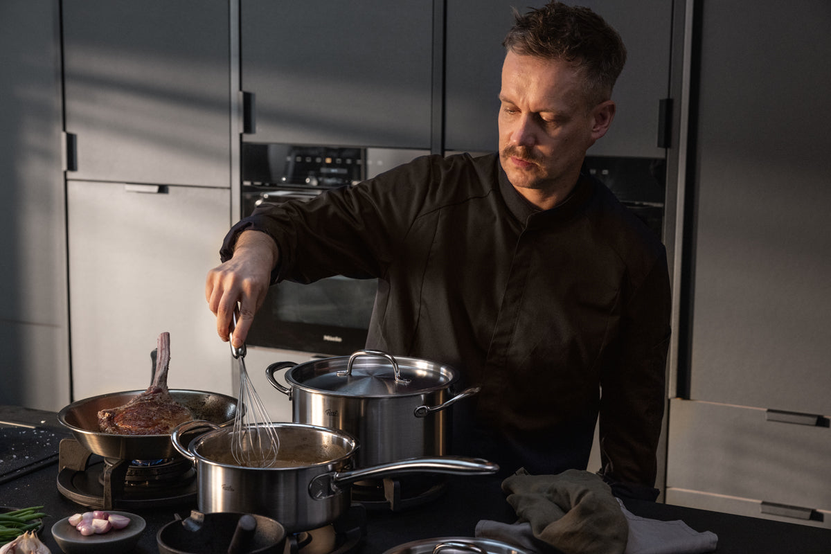 Ein Mann in einem schwarzen Hemd rührt gekonnt in einem hochwertigen Fissler-Edelstahltopf auf dem Herd, daneben steht eine Pfanne aus dem 4-teiligen Fissler M5 Pro-Ply®-Set mit einem Metalldeckel aus recyceltem Edelstahl, umgeben von weiteren hochwertigen Küchenutensilien in einer eleganten, modernen Küche– und demonstriert damit Fisslers Tradition deutscher Präzisionstechnik und zeitlosen Designs.