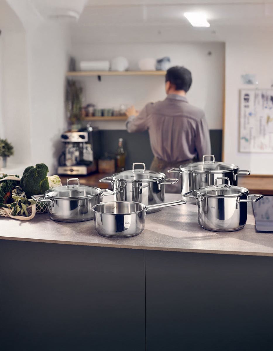 Fissler LumiTherm 5-tlg. Topfset mit Glasdeckel auf moderner Küchenarbeitsplatte neben frischem Gemüse, unscharfe Person im Hintergrund.