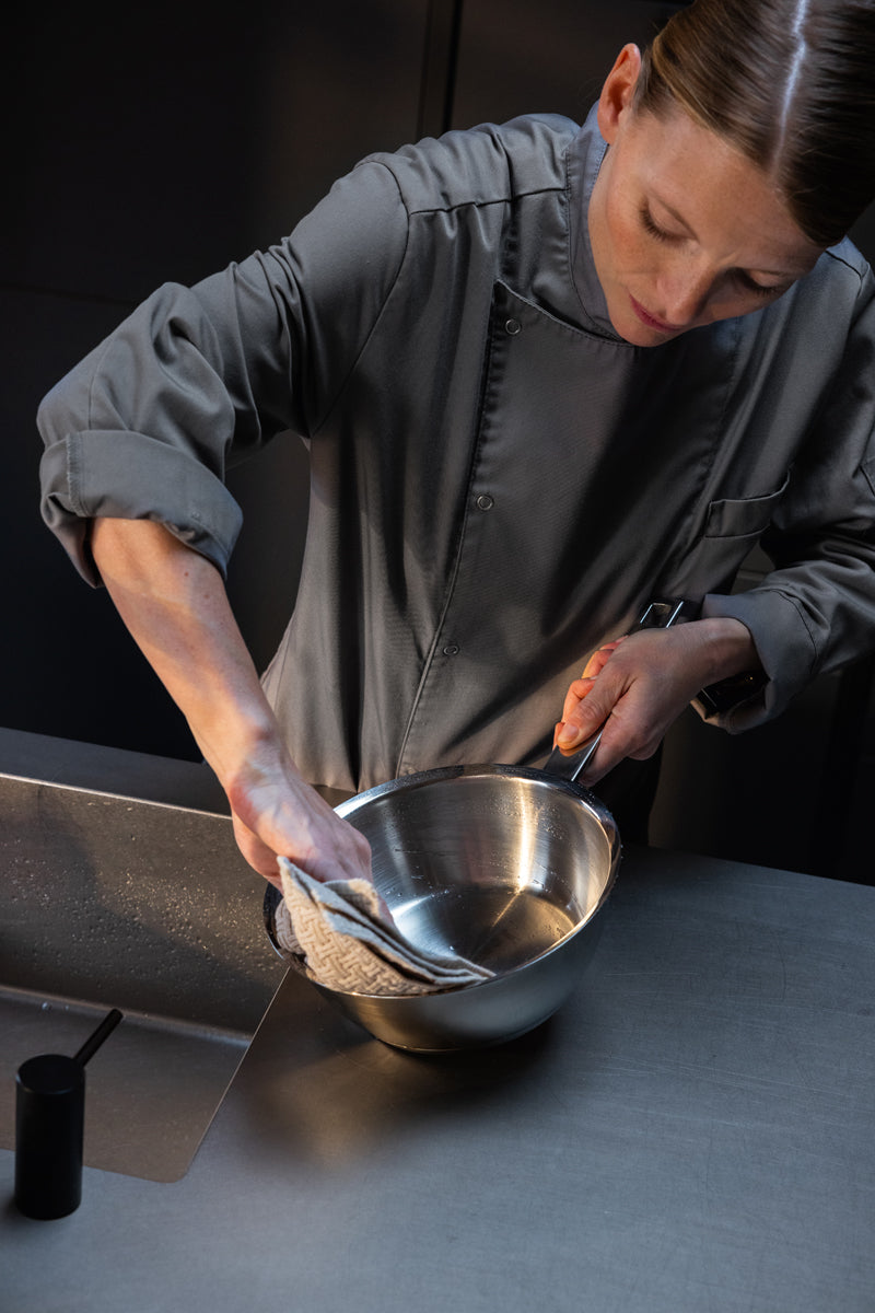 Ein Koch in Grau trocknet das 6-teilige deutsche Edelstahlset der Fissler Original-Profi Collection® mit Glasdeckeln auf einer eleganten Theke.