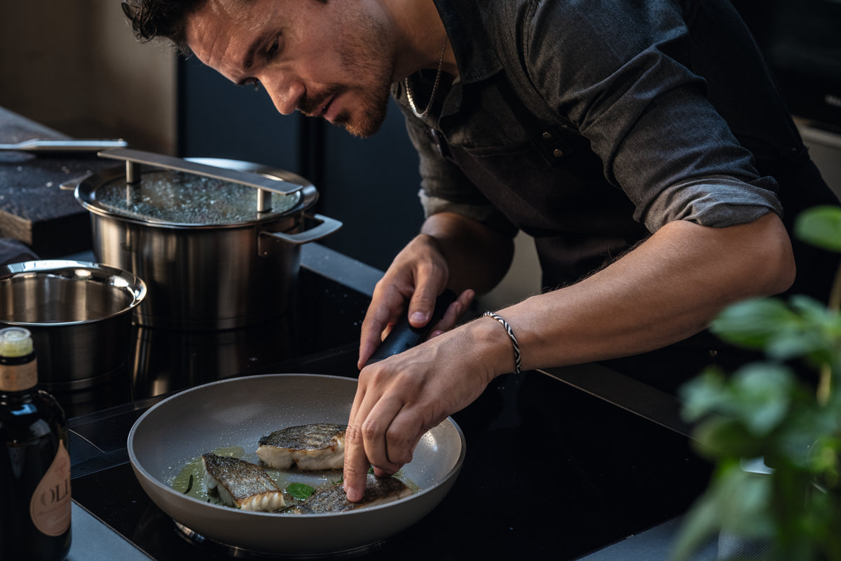 Ein Mann bereitet Fisch mit Kräutern in dem Fissler Ceratal® Evo Classic Pfannenset zu, das deutsche Präzision und zeitloses Design zeigt.