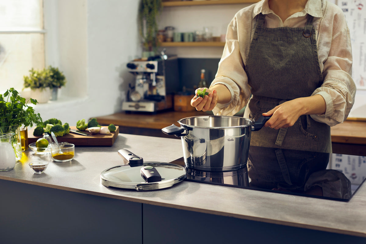 Broccoli held over a Fissler Vitaquick® Elemental pressure cooker in a kitchen, with fresh herbs and the lid on the counter.