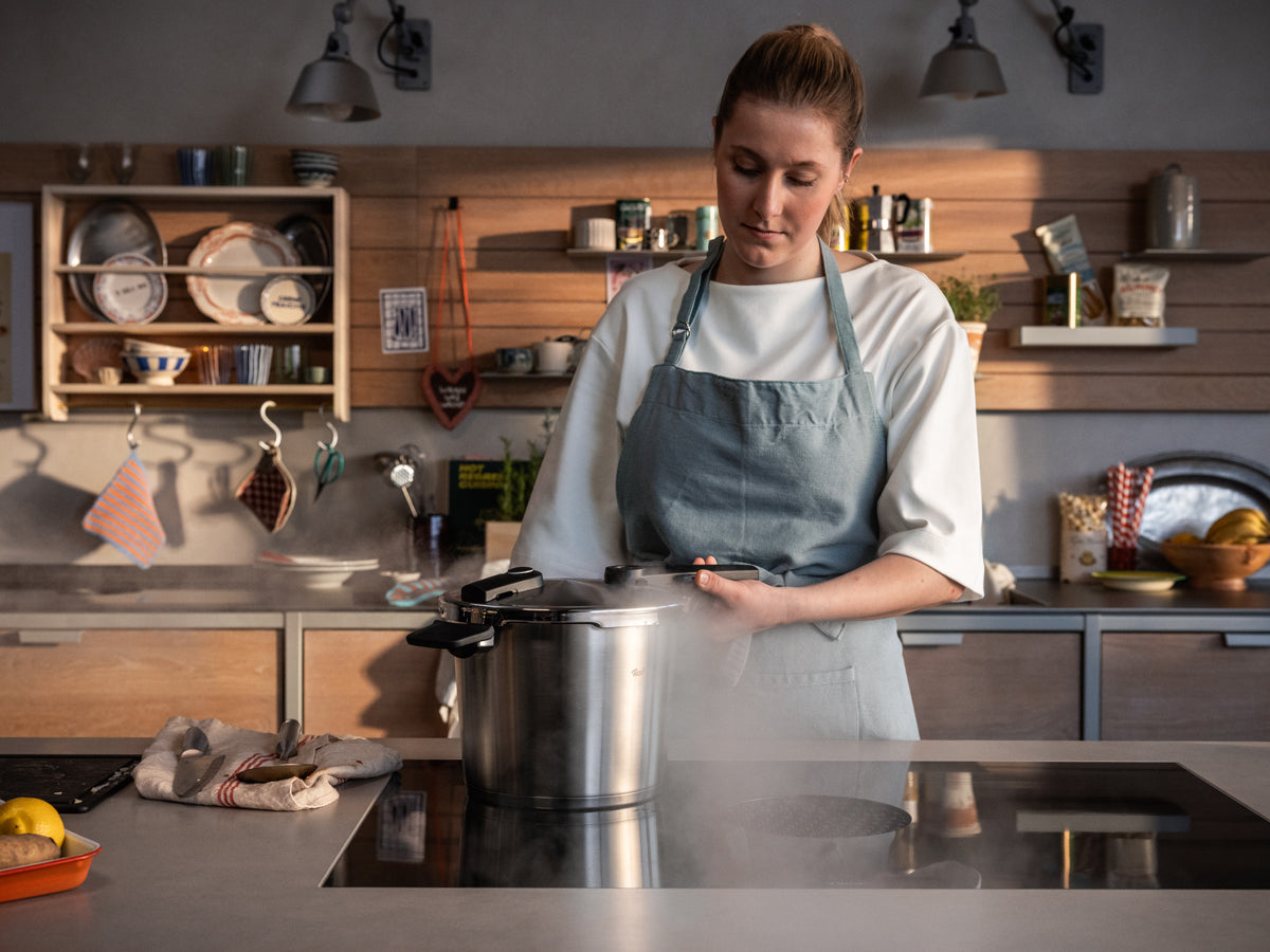 Eine Frau in einer blauen Schürze steht in einer sonnendurchfluteten Küche und lässt vorsichtig Dampf aus ihrem 2-teiligen Fissler Vitaquick® Premium-Schnellkochtopf-Set mit SKT Book auf dem Herd ab. Seit 1845 in Deutschland entworfen und entwickelt, steht unser Kochgeschirr für Präzision, Langlebigkeit und zeitlose Innovation. Regale voller Geschirr und Küchenutensilien bilden eine gemütliche Kulisse, die die elegante Edelstahlkonstruktion dieses hochwertigen Schnellkochtopf-Sets „Made in Germany“ perfekt