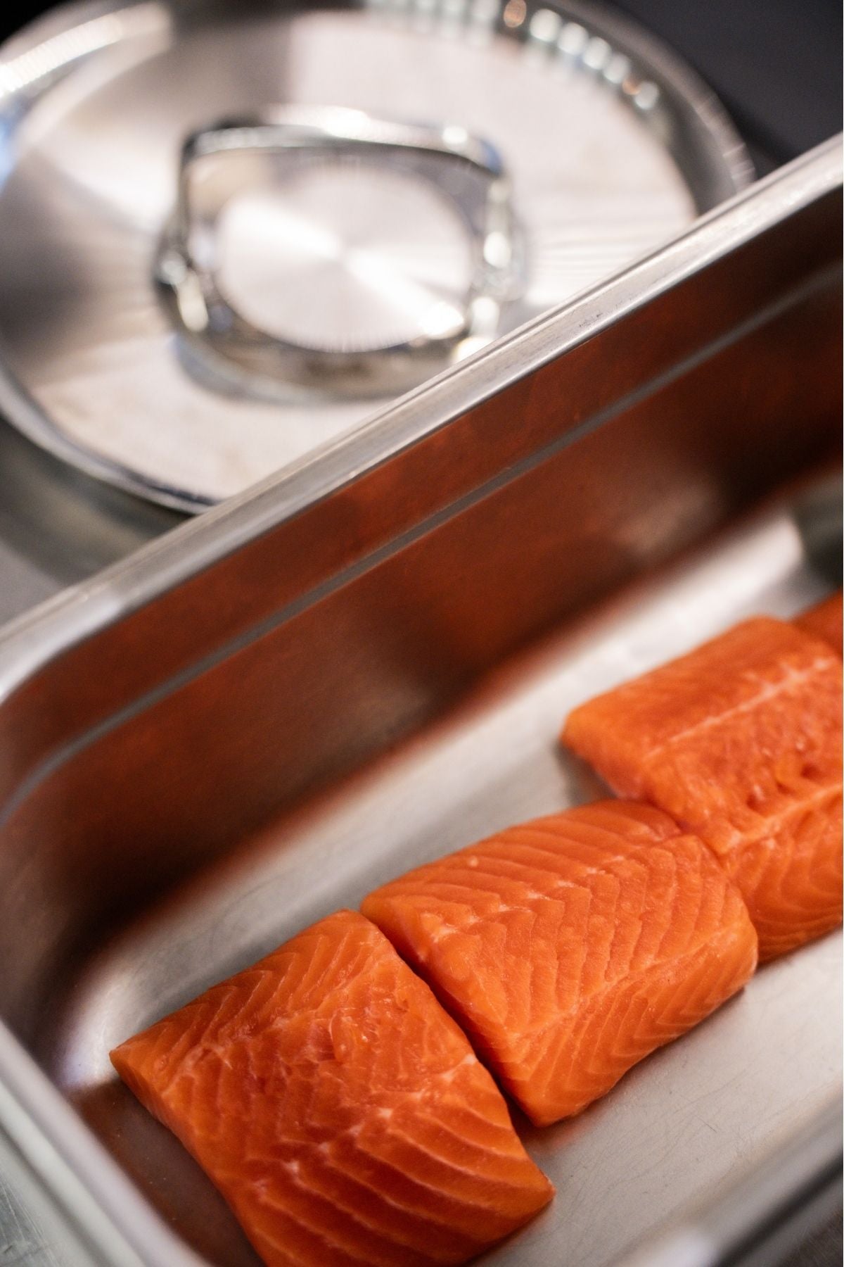 Drei frische Lachsfilets liegen in einem Fissler-Edelstahltablett, hinter dem sich unser ikonischer Topf und Deckel mit deutscher Präzision verbirgt.