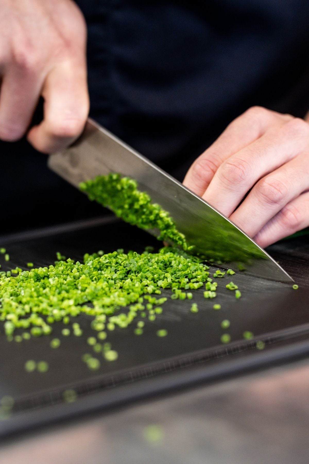 Eine Person hackt frische grüne Kräuter auf einem dunklen Brett und zeigt dabei die Präzision eines hochwertigen Fissler-Küchenmessers.