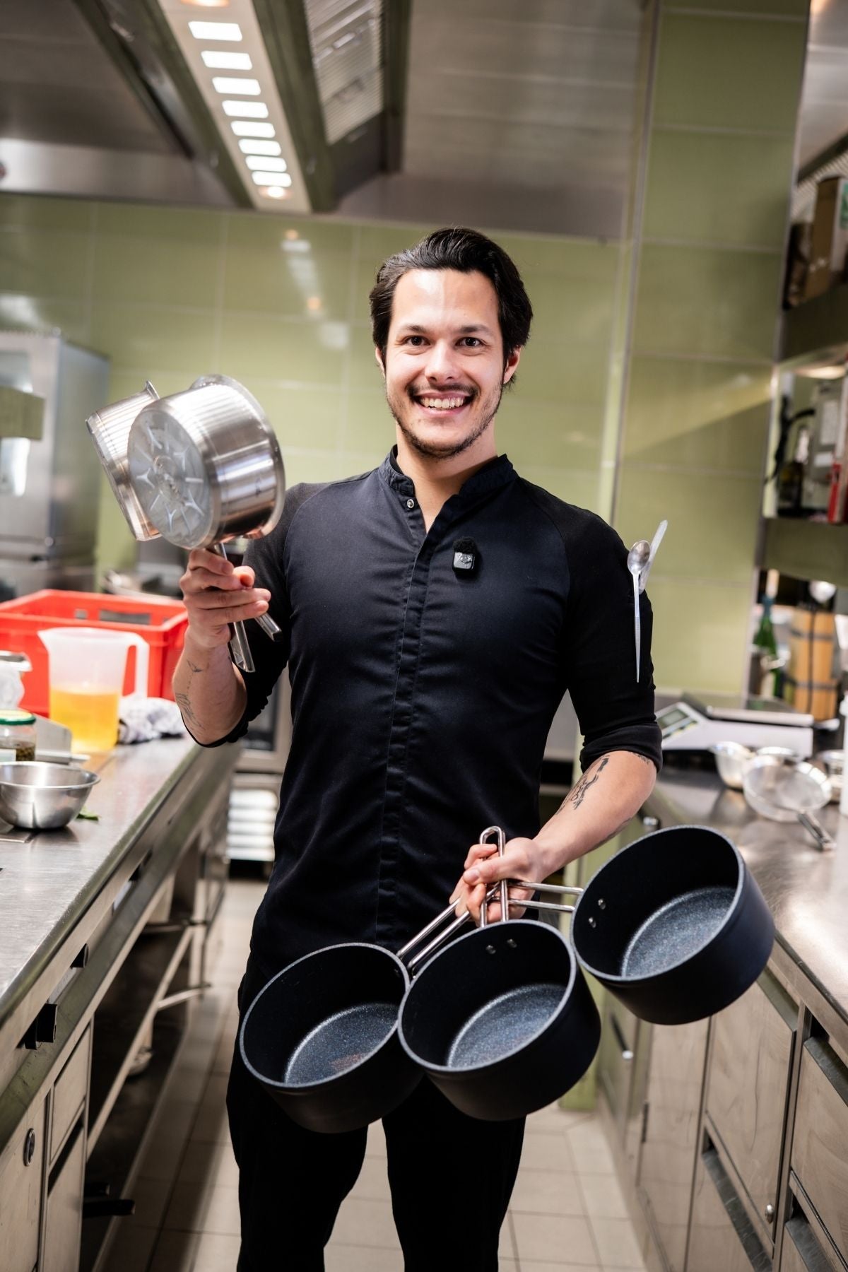 Ein lächelnder Koch in Schwarz hält hochwertiges Fissler-Edelstahl-Kochgeschirr in einer modernen Küche mit grün gekachelten Wänden.
