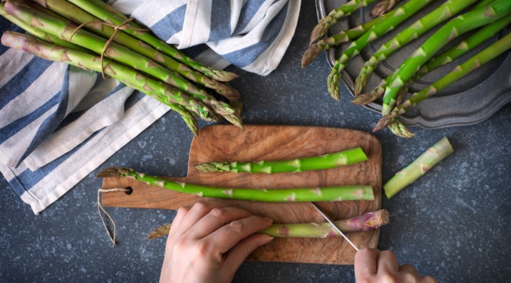 A person cuts green asparagus on a wooden cutting board, with Fissler cookware and kitchen towels on a plain gray counter.