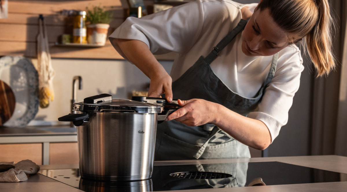 Eine Frau in einer modernen Küche, die eine Schürze trägt, verschließt den Deckel eines hochwertigen Fissler-Edelstahl-Schnellkochtopfs, der auf einem eleganten, modernen Herd steht – ein Beispiel für deutsche Präzisionstechnik und zeitloses Design.