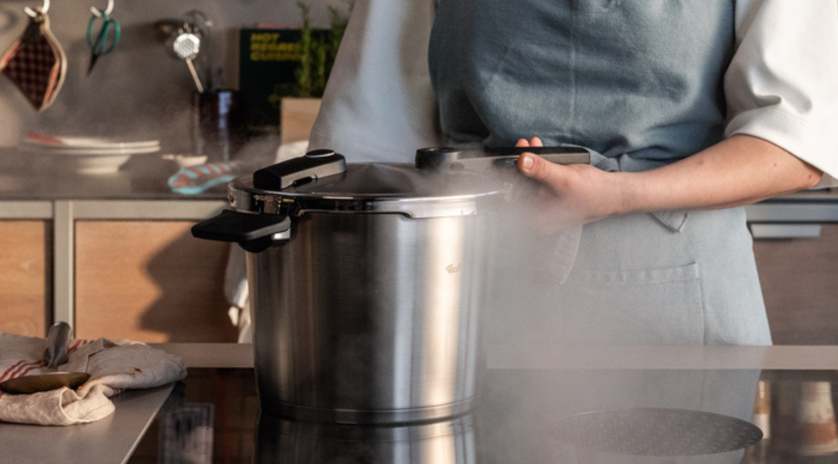 Ein Hobbykoch in einer modernen Küche hebt den Deckel eines dampfenden Fissler-Edelstahl-Schnellkochtopfs und präsentiert damit deutsche Ingenieurskunst und innovatives Design. Der Koch trägt eine Schürze, im Hintergrund sind hochwertige Fissler-Töpfe, -Pfannen und -Zubehörteile ordentlich zwischen frischen Zutaten angeordnet – ein Zeichen für Qualität und Langlebigkeit seit 1845.