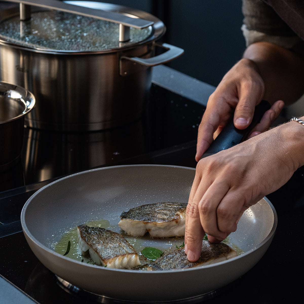 A chef arranges fish fillets in a Fissler pan with oil and herbs; next to it, a covered Fissler pot sits on the stovetop.