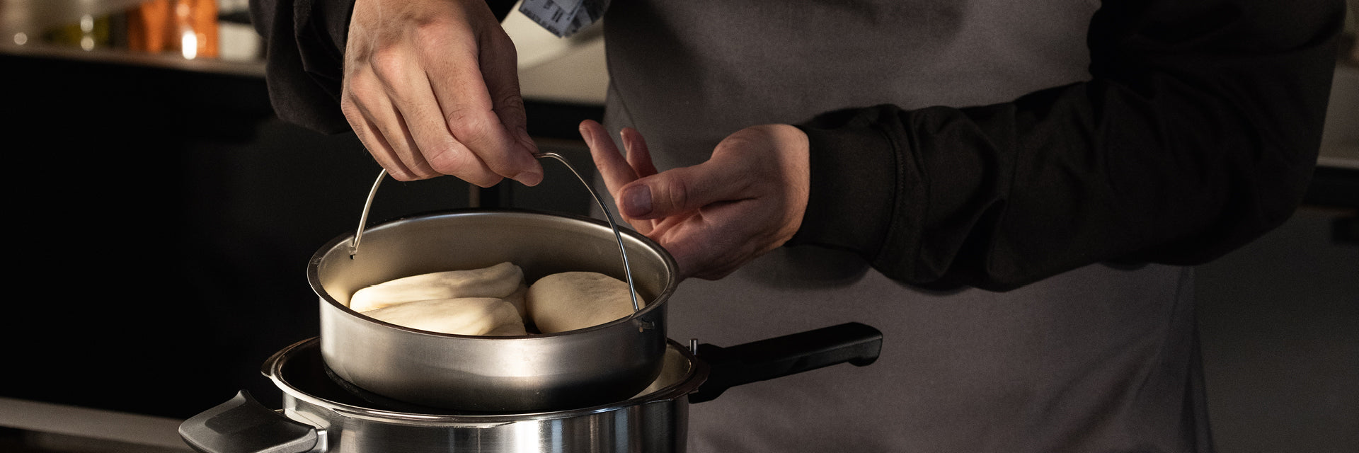 Eine Person in einer grauen Schürze hebt einen Fissler-Edelstahltopf mit Knödeln über einen anderen Topf auf dem Herd, bereit zum Kochen.