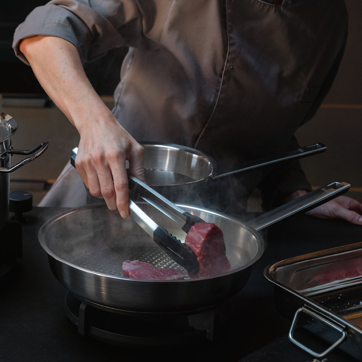 A chef places a raw steak in a Fissler stainless steel pan on the stove, with other high-quality Fissler cookware nearby.