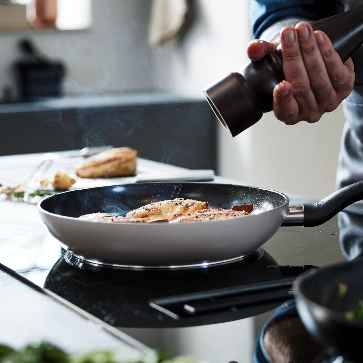 A person seasons fish fillets cooking in a high-quality Fissler stainless steel pan, with steam rising in a modern kitchen.