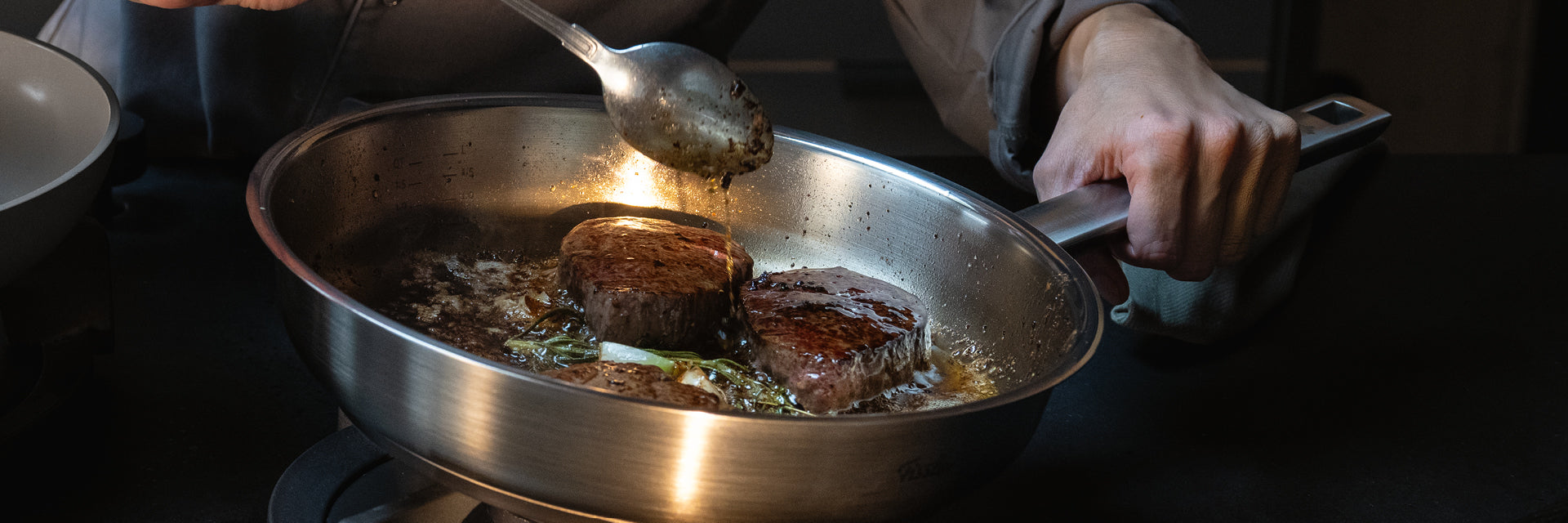 Ein Koch brät zwei Steaks in einer Fissler-Edelstahlpfanne an und begießt sie mit Butter und Kräutern, um ein perfektes Ergebnis auf deutschem Kochgeschirr zu erzielen.