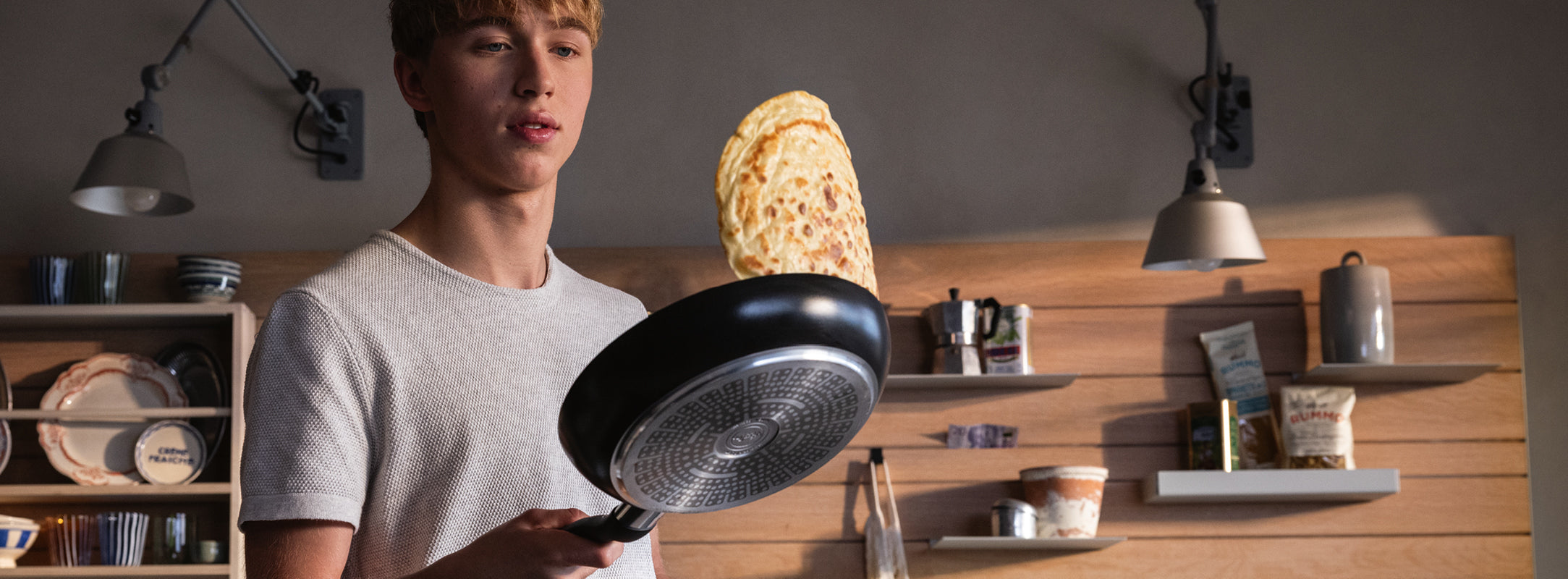Ein junger Mann wendet eine Crêpe in einer Fissler-Pfanne in einer modernen Küche mit Holzschränken, in der deutsches Qualitätskochgeschirr präsentiert wird.