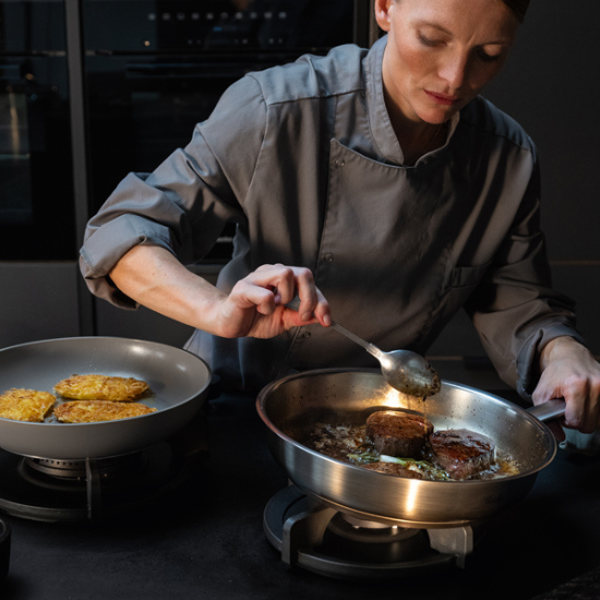 A chef prepares filet mignon in a high-quality Fissler pan, while golden potato fritters cook in another Fissler pan on the stove.
