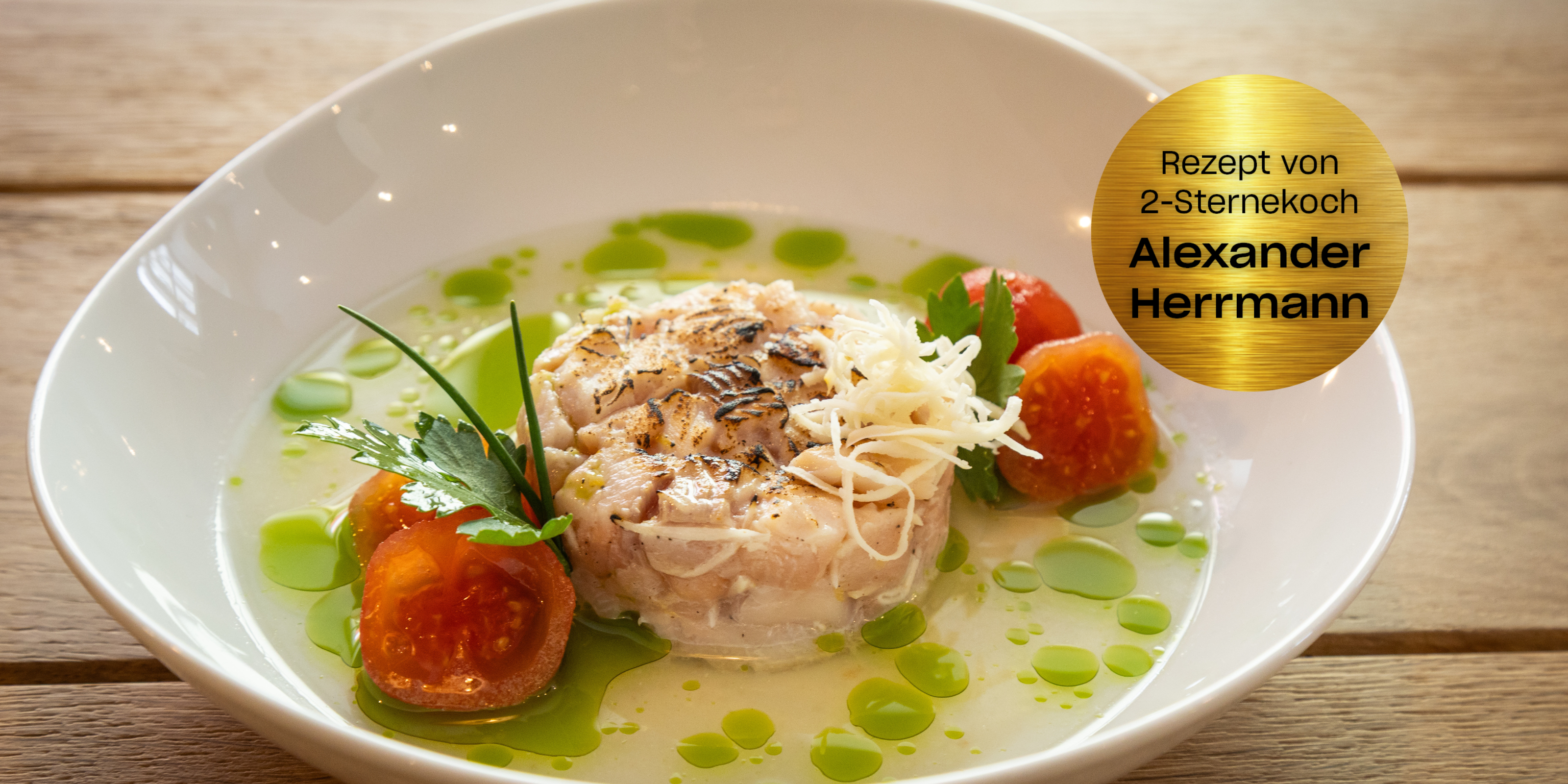 Ein Gourmet-Fischgericht mit Käse, Tomaten, Petersilie und grünem Öl in einer weißen Schale. Goldenes Etikett: Rezept von 2-Sternekoch Alexander Herrmann.
