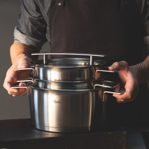 Eine Person in einer dunklen Schürze hält zwei gestapelte Fissler-Edelstahltöpfe mit stabilen Griffen und Deckeln, die auf einer glatten dunklen Oberfläche präsentiert werden.