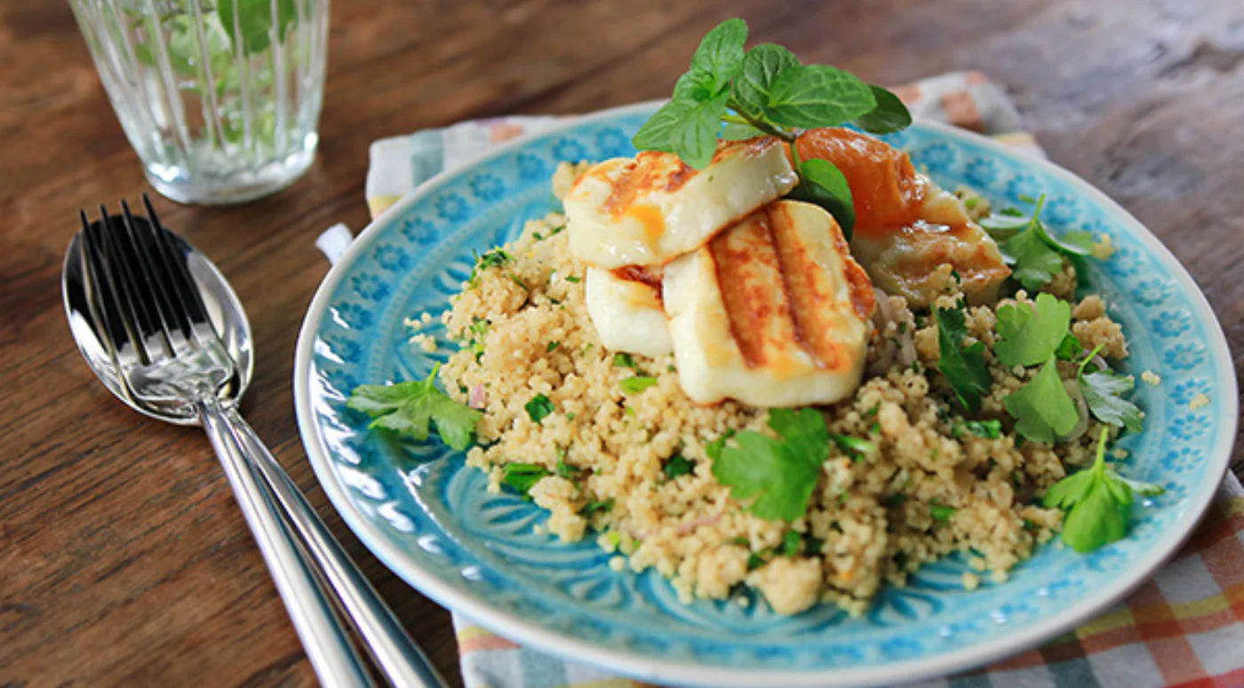 A beautifully arranged blue plate of couscous with grilled halloumi, fresh herbs, and roasted tomatoes sits atop a wooden table, accompanied by a fork and spoon—crafted with Fissler’s renowned stainless steel quality—and a clear glass of water. The elegant setup highlights precision design and timeless style that reflects Fissler’s heritage in premium German cookware.