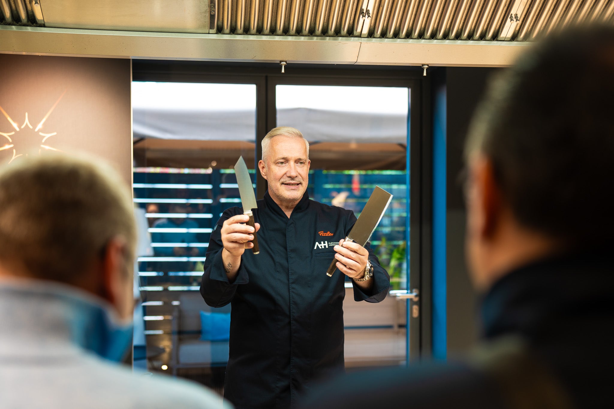 A chef in black holds two knives and presents high-quality Fissler cookware to the audience in a modern kitchen setting.