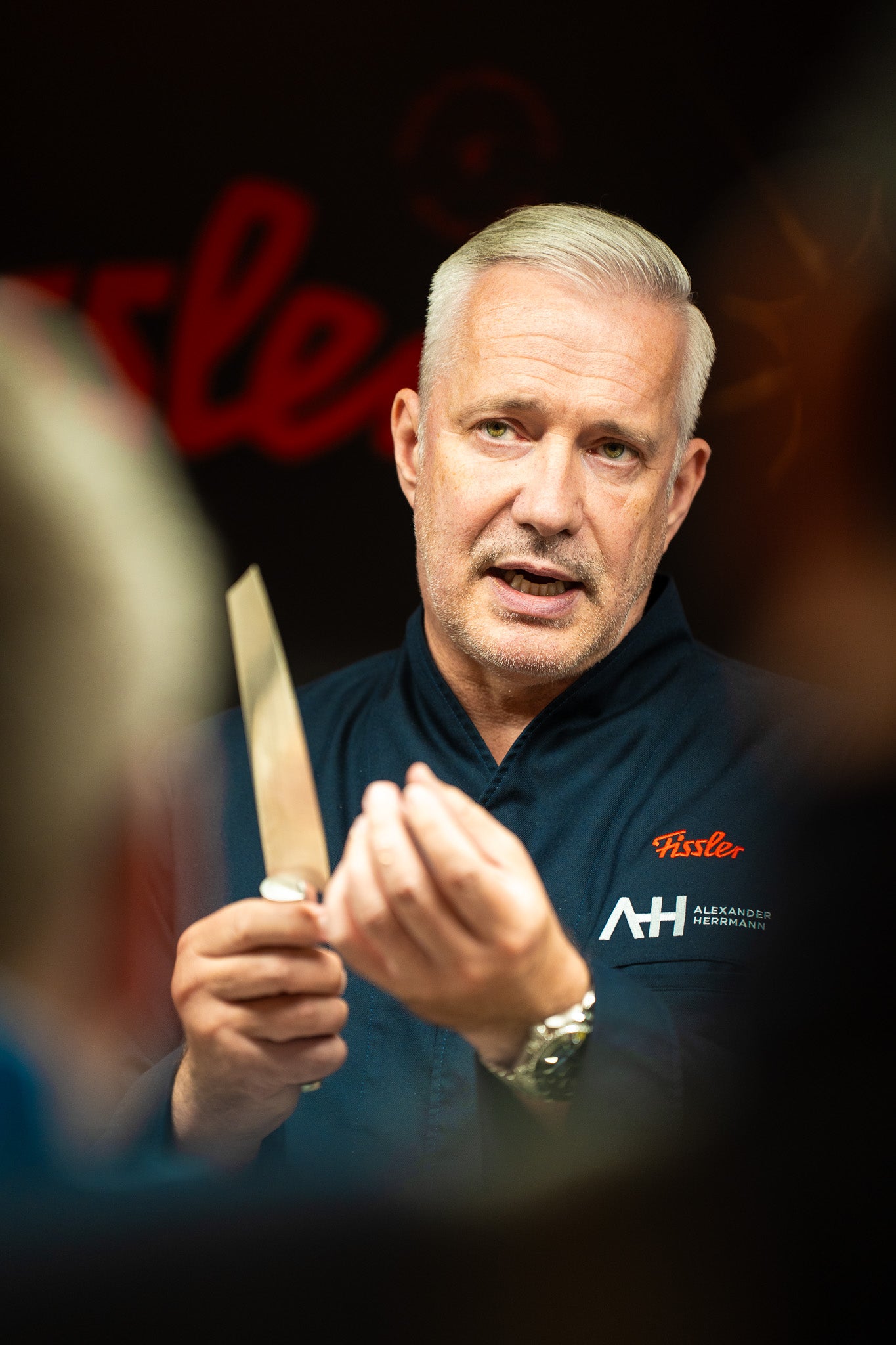 A chef in a dark blue jacket holds up a Fissler knife and gestures while speaking. Blurred background with red Fissler branding.
