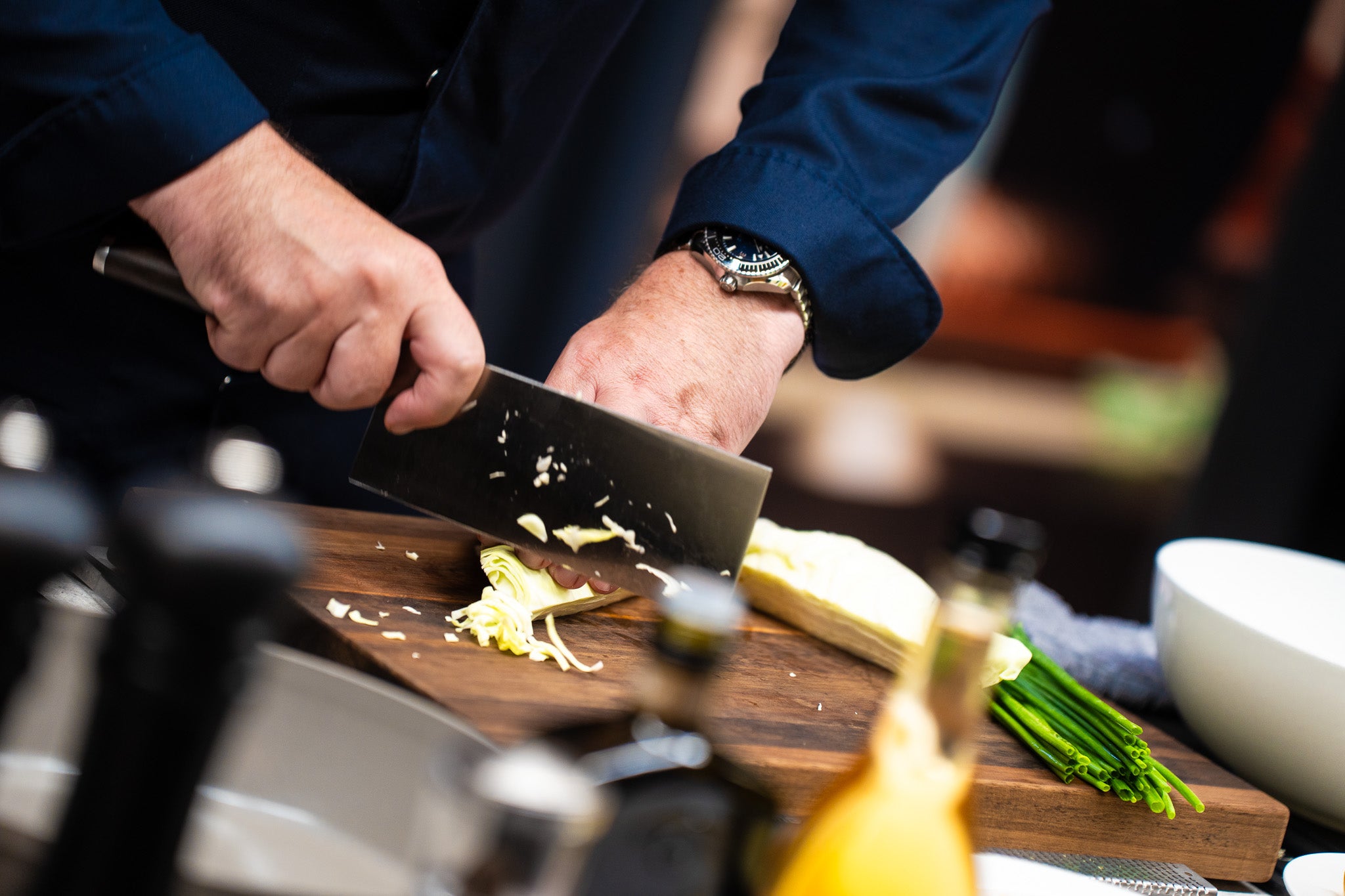 A person in a dark blue shirt cuts vegetables with a Fissler knife on a wooden cutting board, with Fissler cookware and green onions next to it.