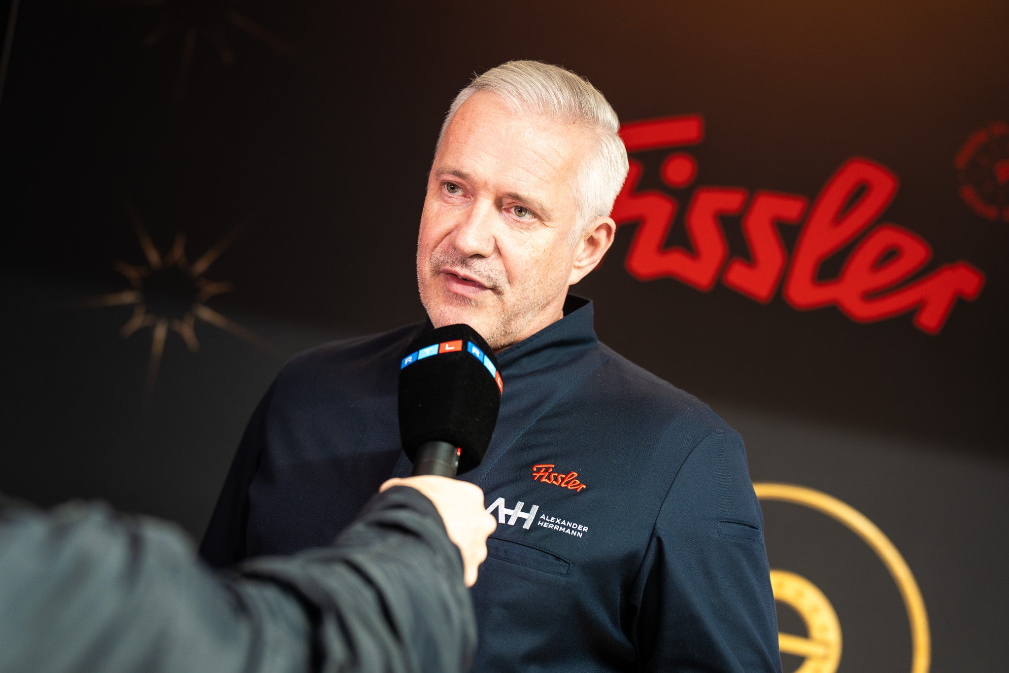 A man in a Fissler jacket is interviewed, with the iconic red logo behind him, representing innovative German cookware since 1845.