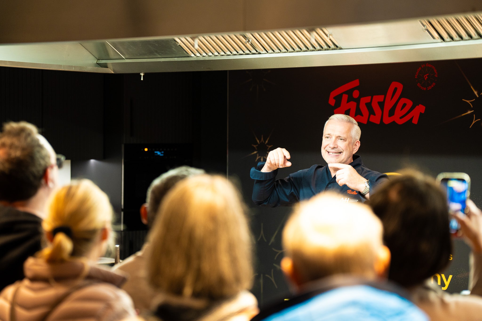 A smiling man stands in front of a Fissler sign, presenting to the audience while guests focus on the high-quality cookware.