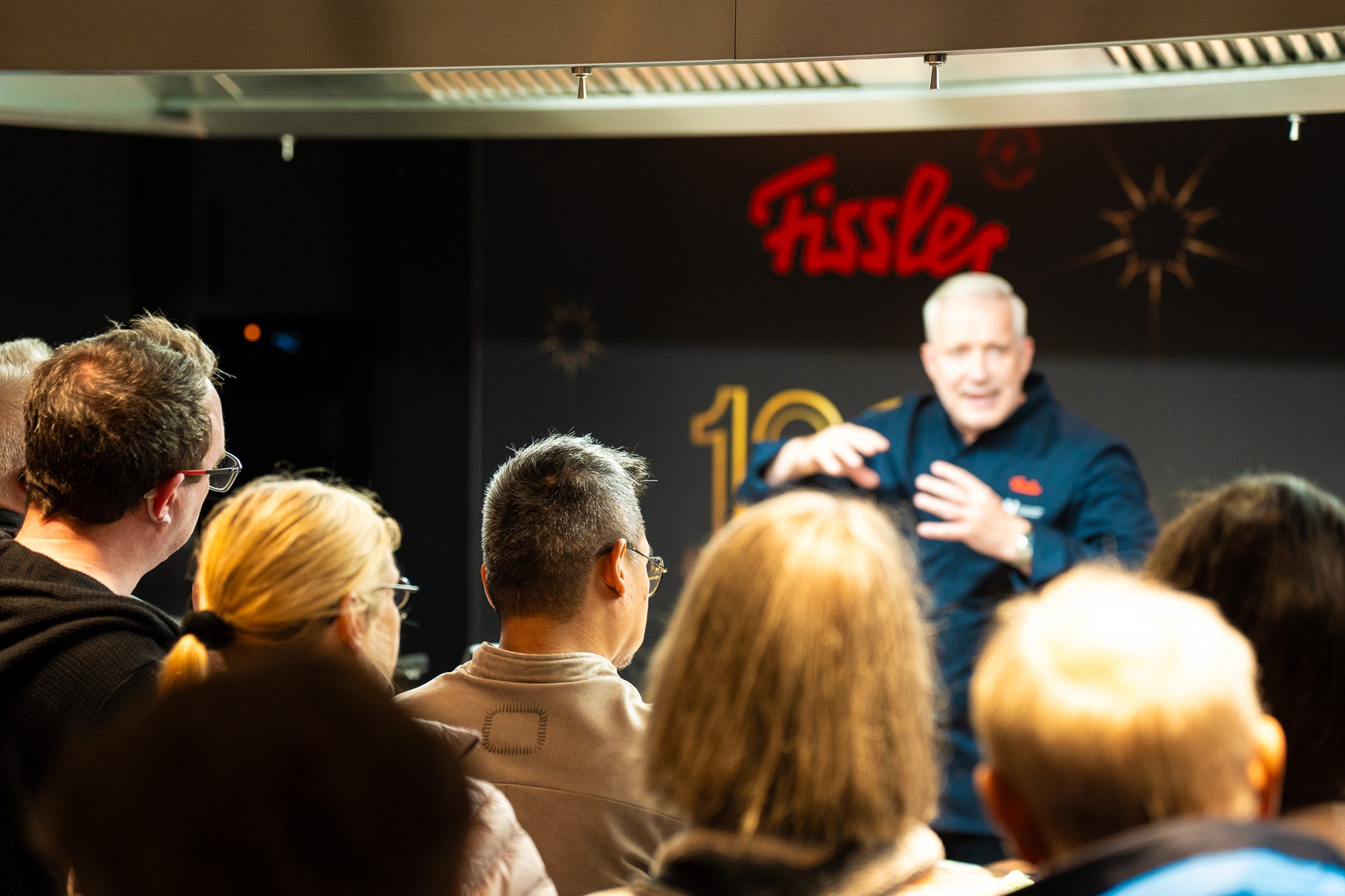 A head chef presents to the audience in a modern kitchen, with a Fissler sign and cookware, representing German precision since 1845.